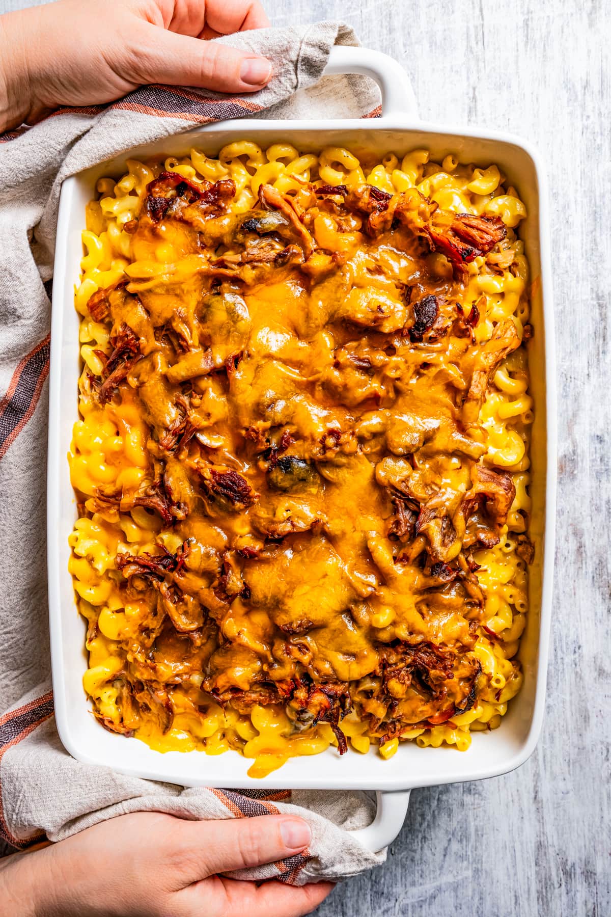 Two hands holding either side of a casserole dish full of pulled pork mac and cheese.
