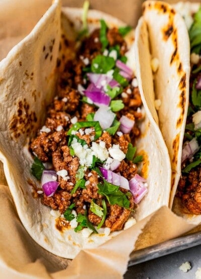 Close-up photo of a taco stuffed with taco meat, cilantro, and red onions on a tray lined with parchment paper.