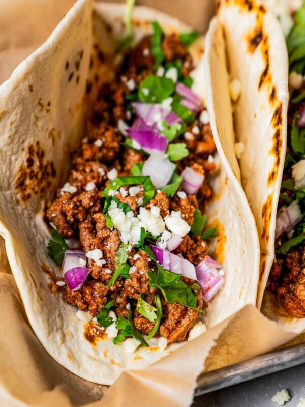 Close-up photo of a taco stuffed with taco meat, cilantro, and red onions on a tray lined with parchment paper.