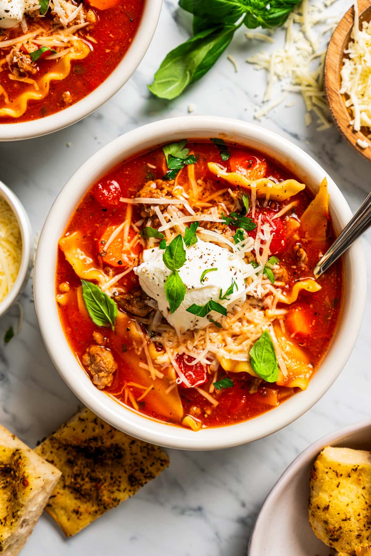 Overhead image of crockpot lasagna soup in a bowl topped with ricotta near a bowl of grated parmesan.