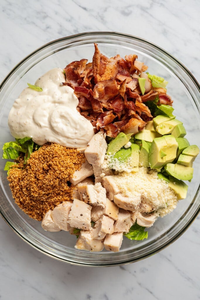 A mixing bowl with bacon, cubed chicken, avocado, cheeses, and dressing to make a filling for tortilla wraps.