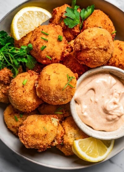 Close-up photo of potato croquettes served in a bowl with a side of aioli and lemon wedges.