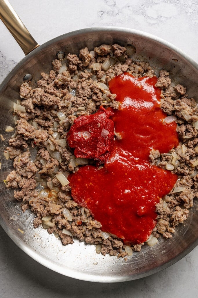 Adding tomato sauce, crushed tomatoes, and tomato paste to browned, seasoned meat.