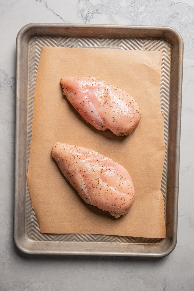 2 raw seasoned chicken breasts on a sheet pan.