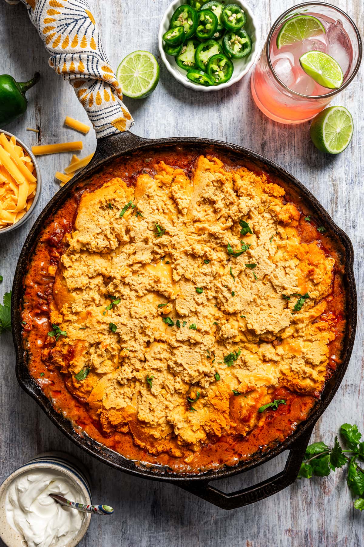 Overhead image of tamale pie in a skillet near jalapenos, sour cream, and cheddar cheese.