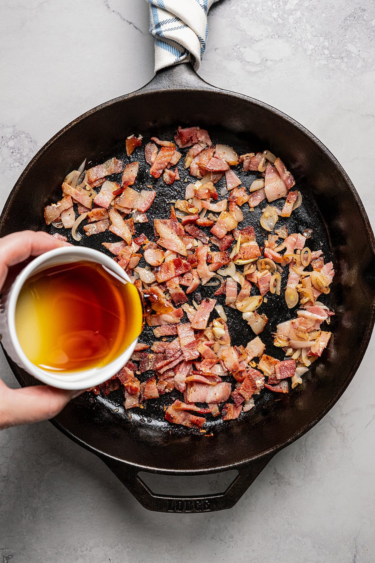 Maple syrup being poured from a white ramekin over diced bacon in a black skillet.