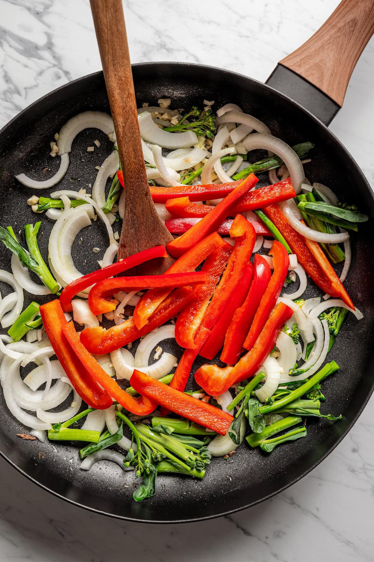 Red pepper slices, onions, and broccolini stems in a skillet with a wooden spoon.