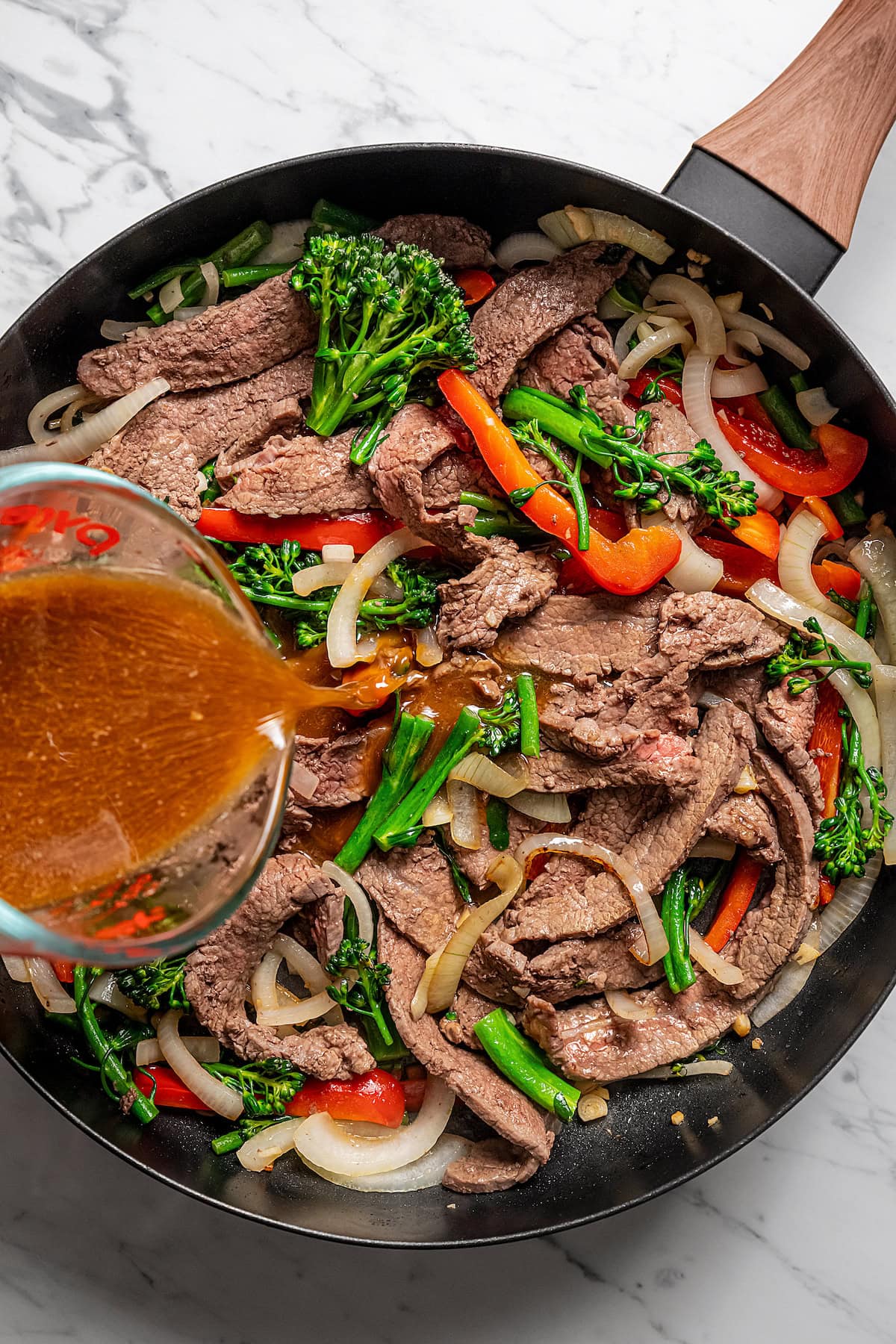 Stir fry sauce is poured over sauteed beef, red peppers, and broccolini in a skillet.
