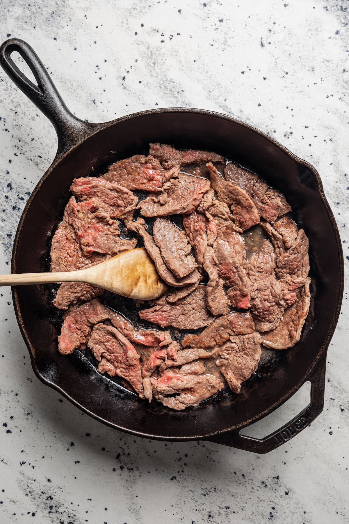 A wooden spoon stirring flank steak slices in a skillet.