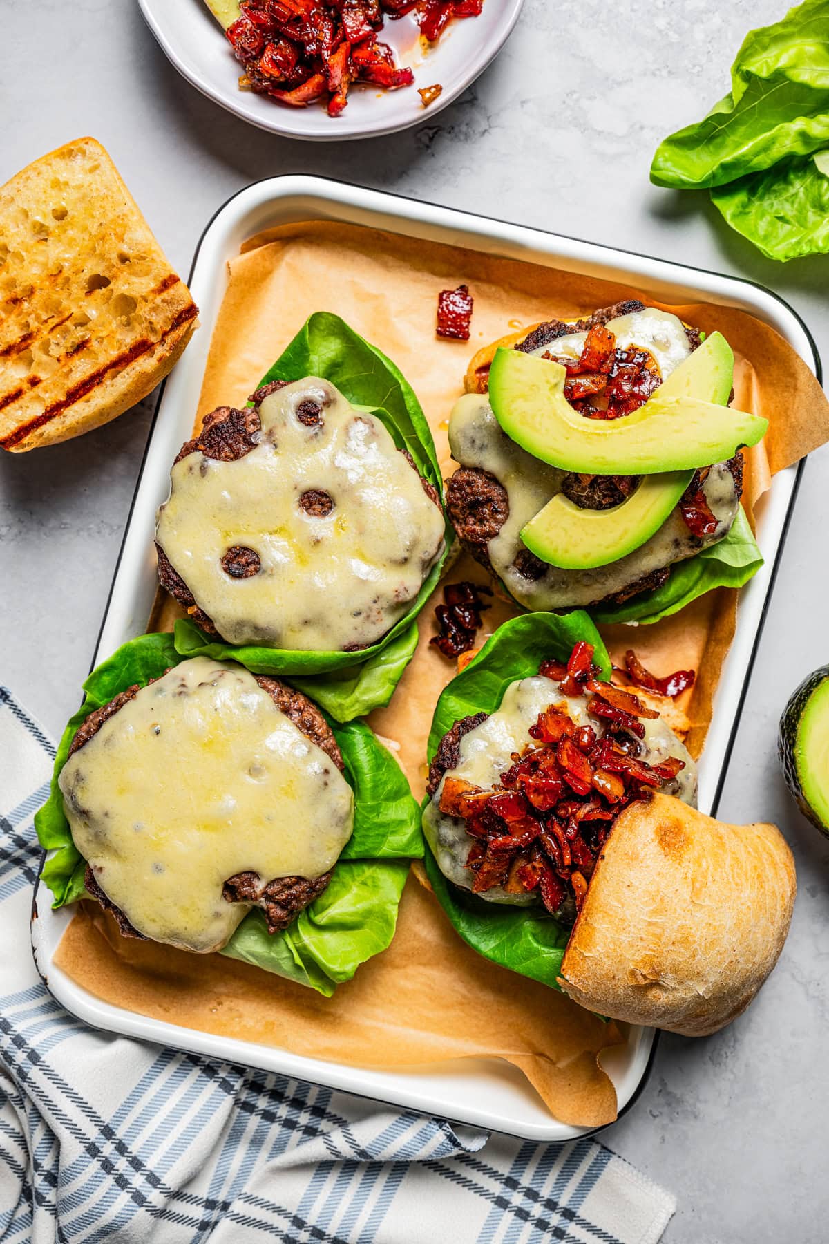 Overhead view of partially assembled California burgers topped with lettuce, cheese, avocado slices, and bacon jam on a lined metal tray, surrounded by toppings.