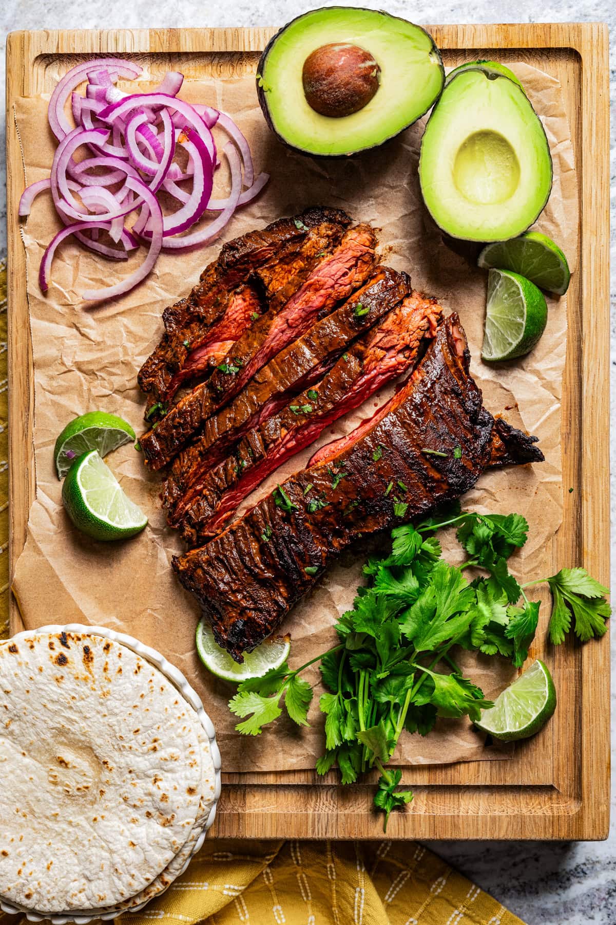 Sliced garlic guajillo steak next to cilantro, sliced onions, lime wedges, and half of an avocado on a wooden cutting board.
