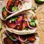 Overhead view of three garlic guajillo steak tacos garnished with sliced onions and cilantro, next to scattered lime wedges.