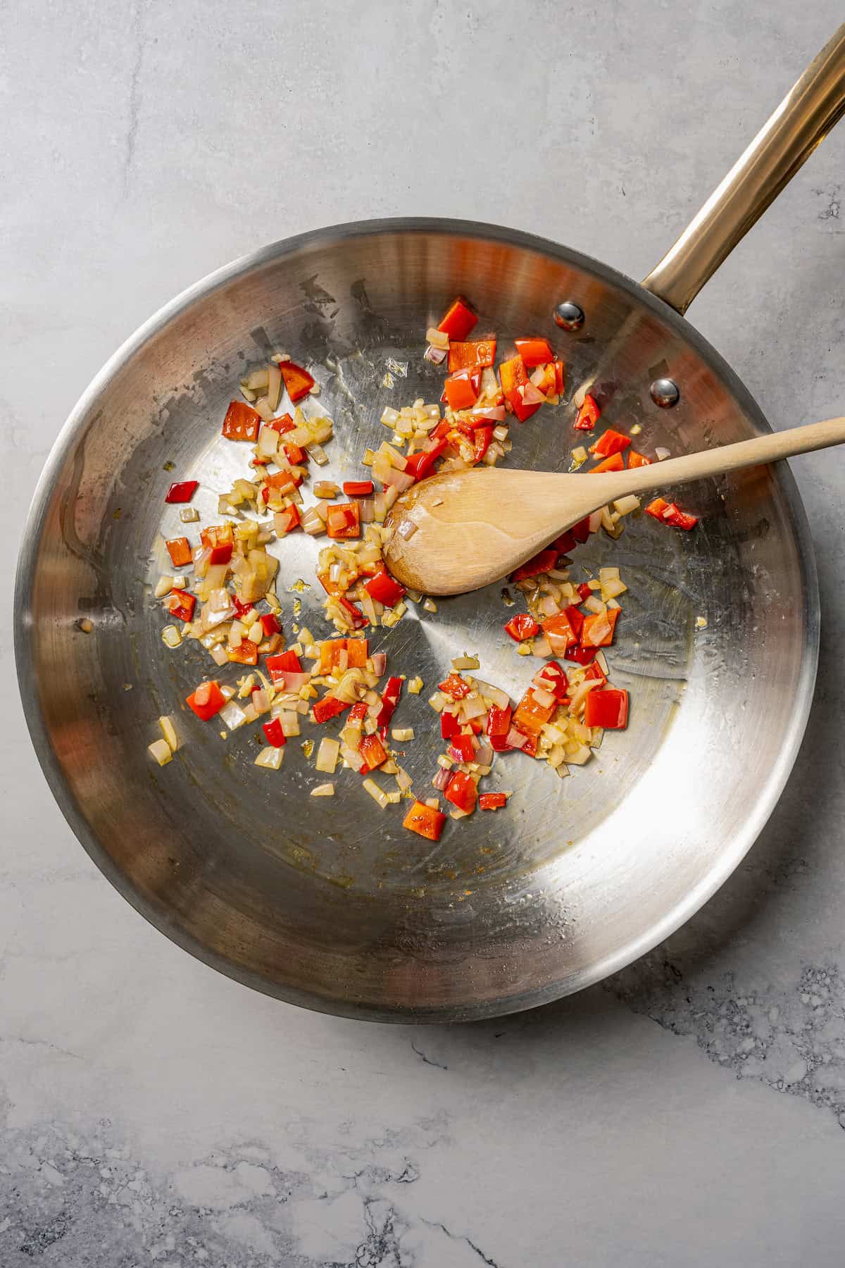 Diced red bell peppers and onion sautéing in a skillet with a wooden spoon.