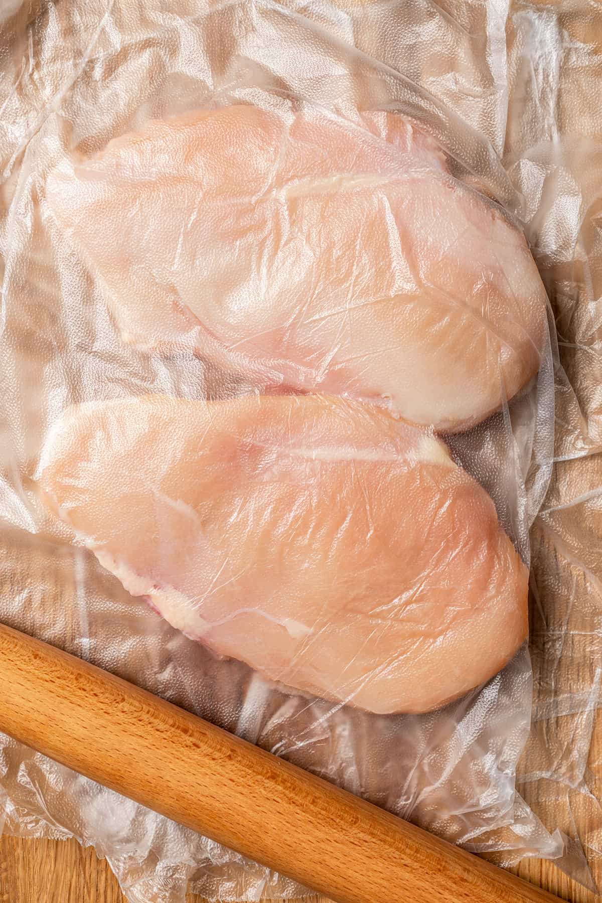 Two chicken breasts pounded thin between two sheets of plastic wrap, next to a wooden rolling pin on a wooden cutting board.