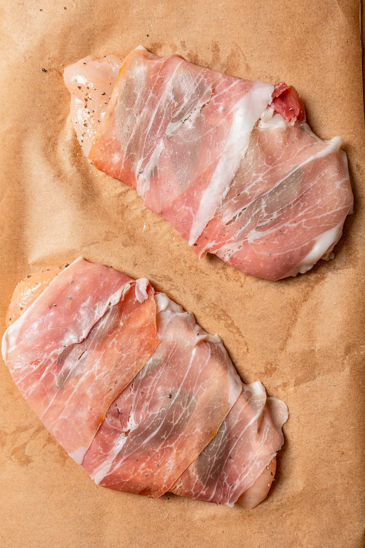 Overhead view of two chicken breasts wrapped in prosciutto on a piece of parchment paper.