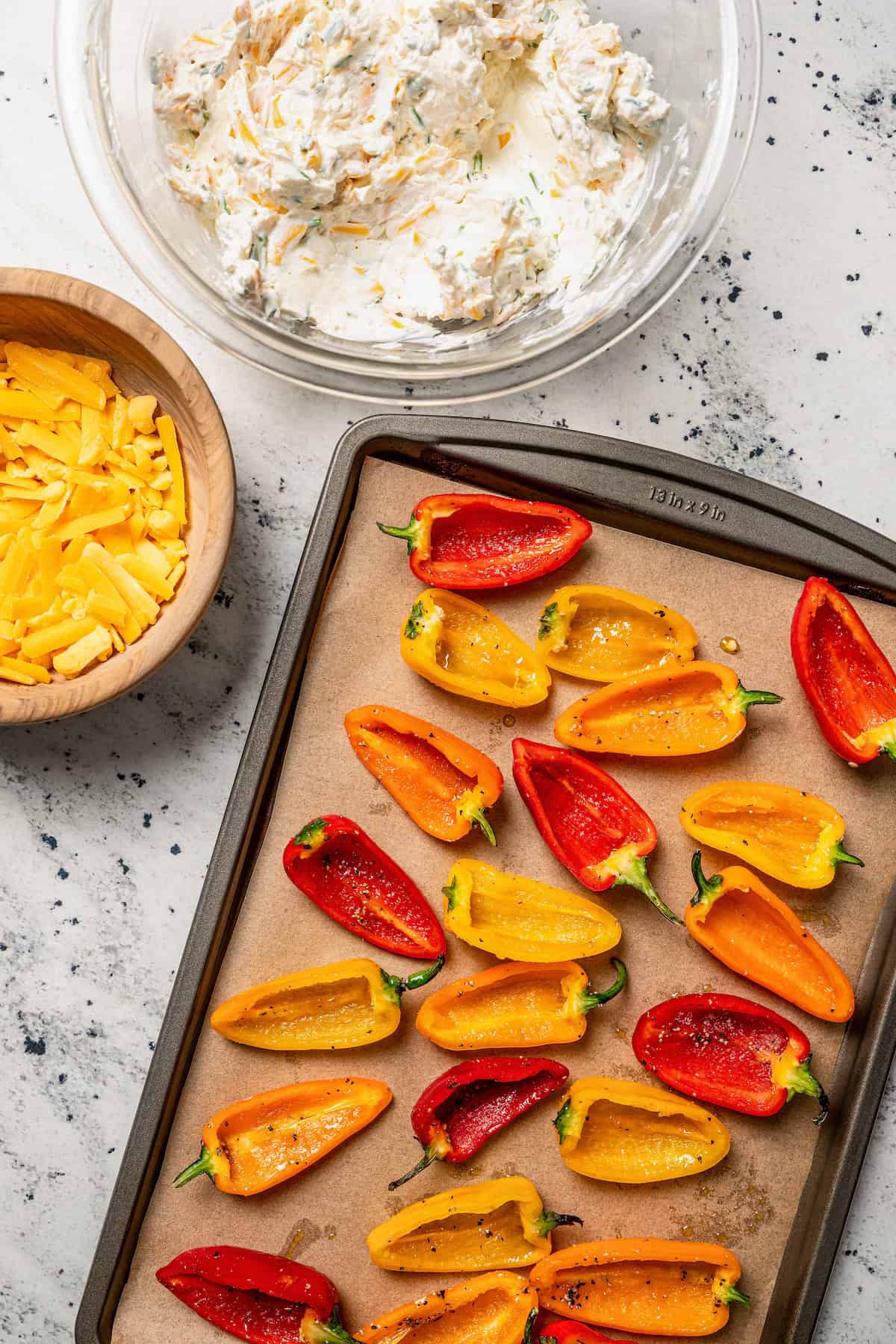 Par-baked bell pepper halves on a lined baking sheet, next to bowls of cream cheese filling and shredded cheddar cheese.