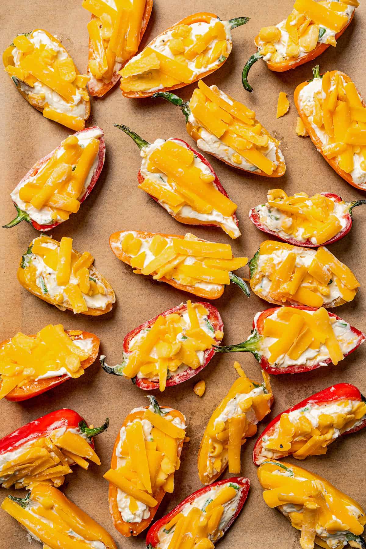 Mini bell peppers stuffed with cream cheese and topped with shredded cheese on a lined baking sheet.