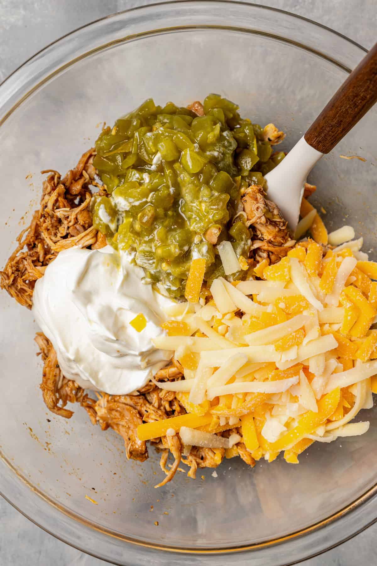 Green chili chicken enchilada filling ingredients combined in a glass bowl with a spatula for stirring.
