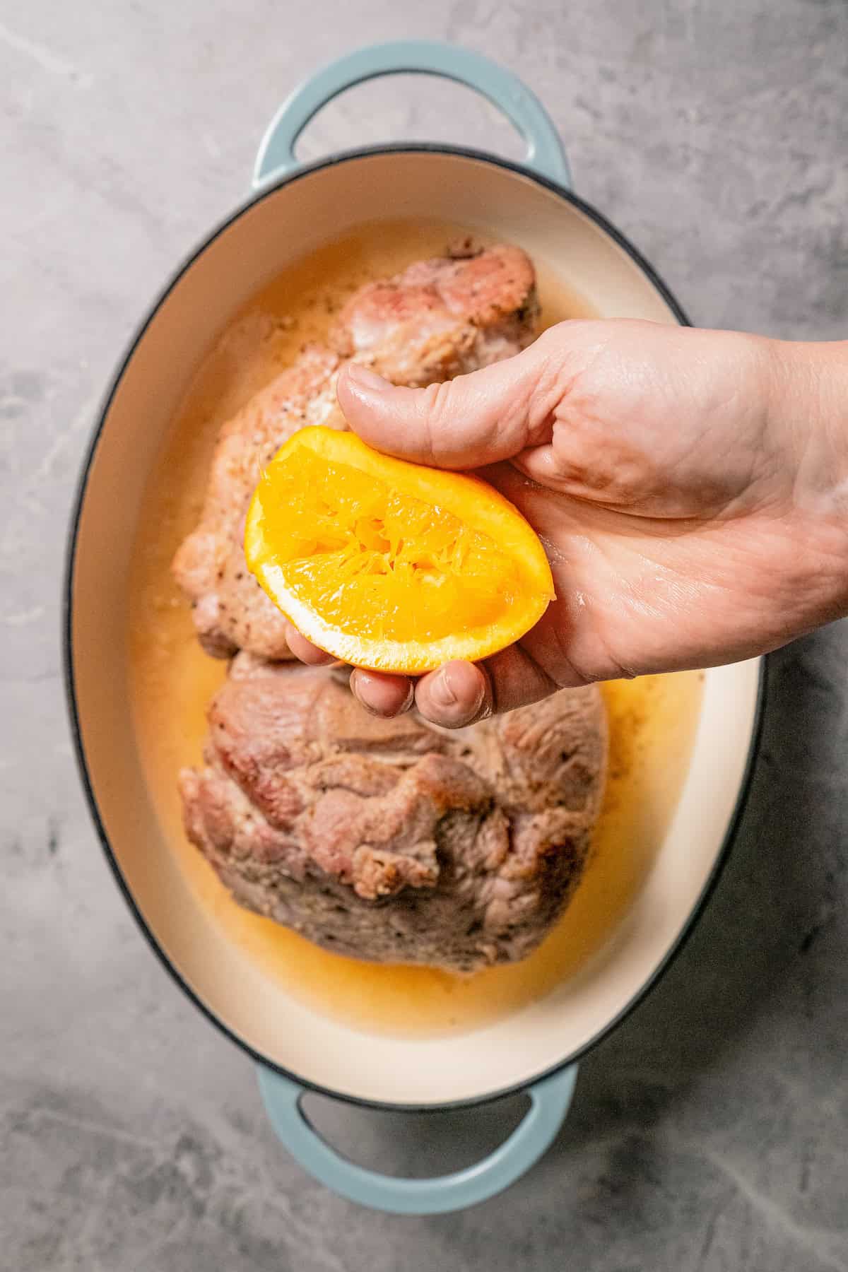 Overhead view of a hand holding a freshly squeezed orange over a pork roast in a baking dish.