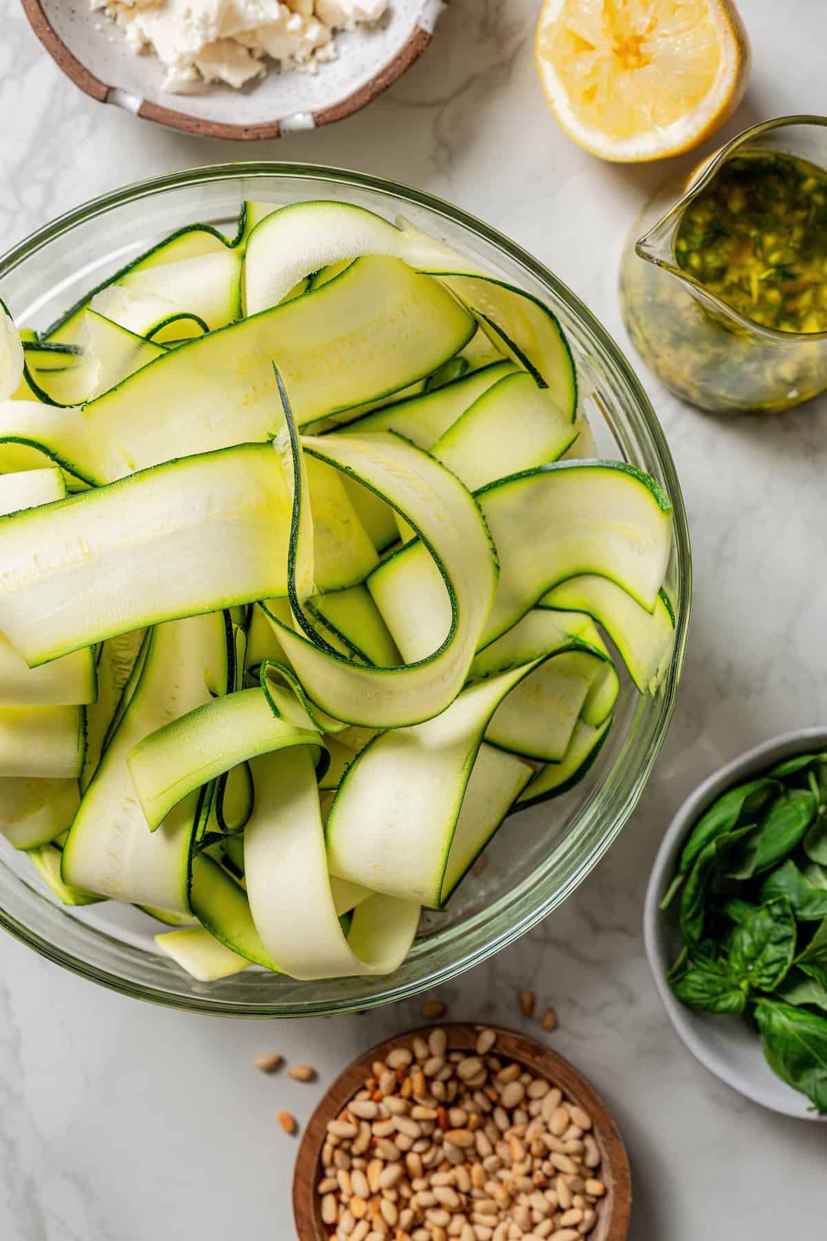 Zucchini ribbons in a glass bowl surrounded by smaller bowls filled with salad ingredients.