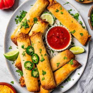 Flautas arranged on a serving platter with a bowl of dipping sauce. Slice jalapenos, lime wedges, tomatoes, and cheese are placed near the platter.