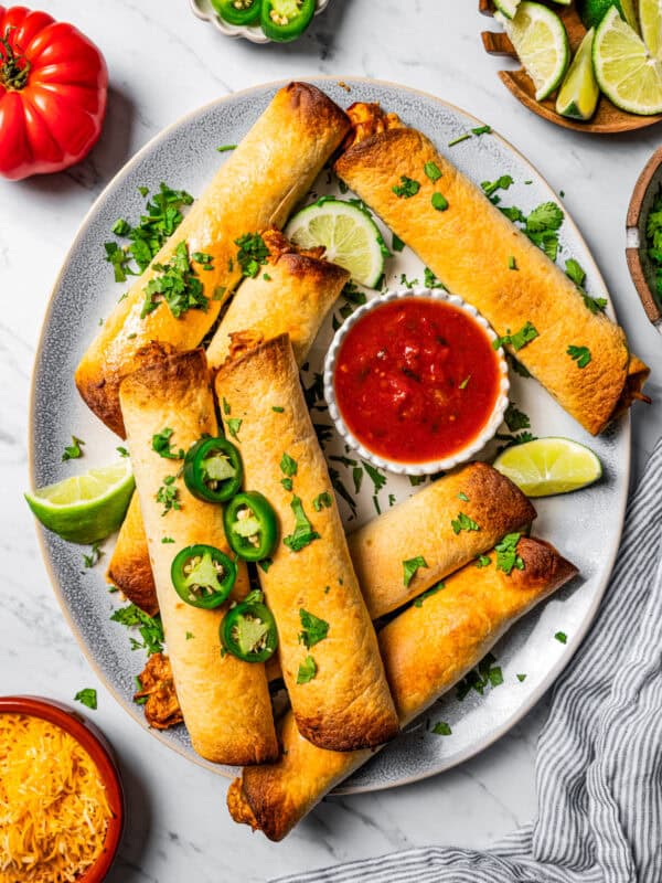 Flautas arranged on a serving platter with a bowl of dipping sauce. Slice jalapenos, lime wedges, tomatoes, and cheese are placed near the platter.