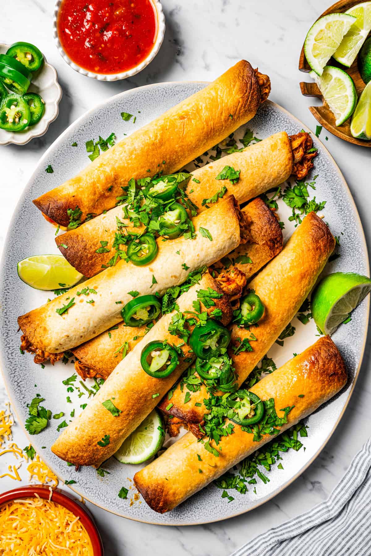 Overhead view of crispy chicken flautas on a round platter, garnished with lime wedges, sliced jalapeños, and fresh cilantro, surrounded by small bowls with more garnishes.