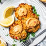 Overhead view of three air fried crab cakes topped with remoulade and garnished with lemon wedges on a white plate, next to a fork.