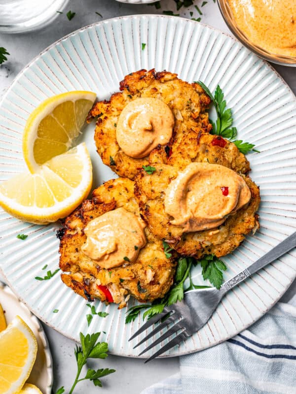 Overhead view of three air fried crab cakes topped with remoulade and garnished with lemon wedges on a white plate, next to a fork.