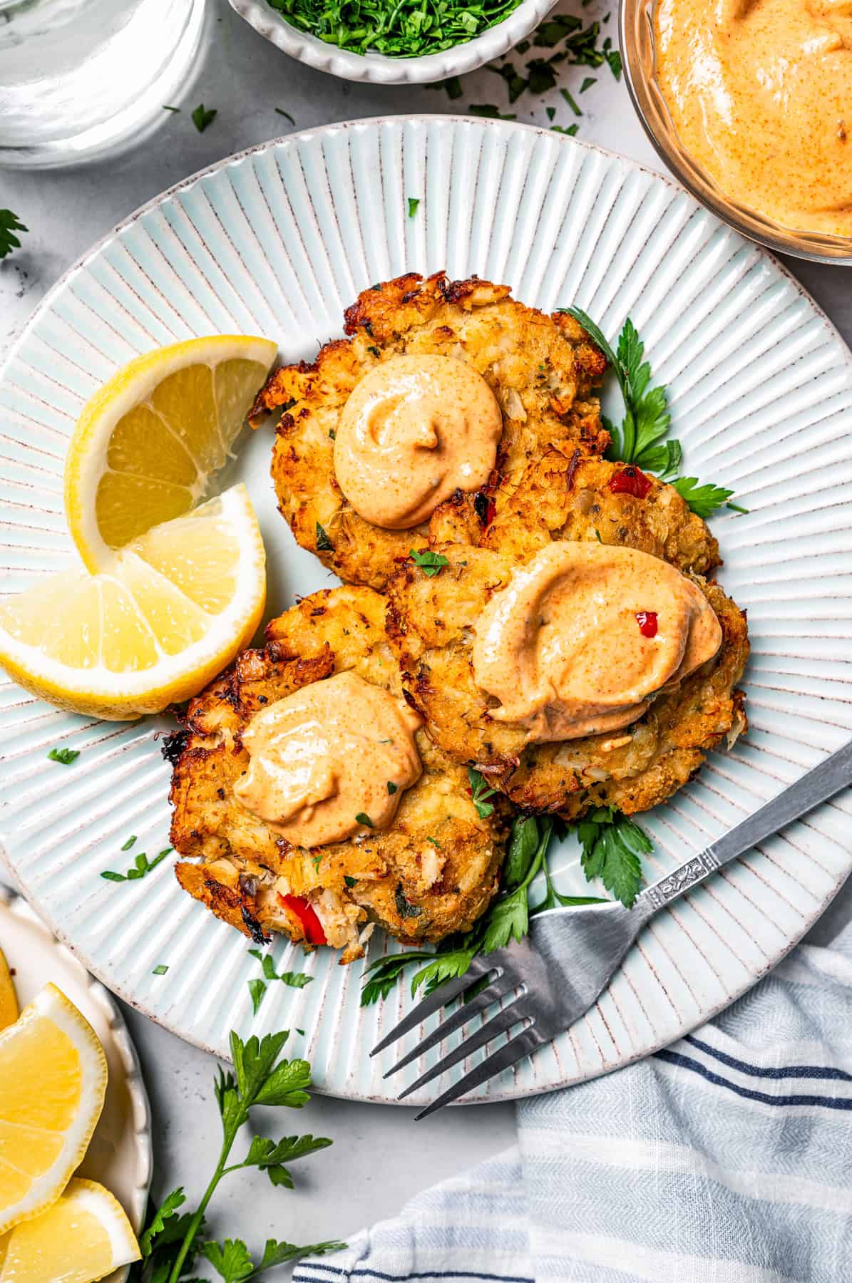Overhead view of three air fried crab cakes topped with remoulade and garnished with lemon wedges on a white plate, next to a fork.
