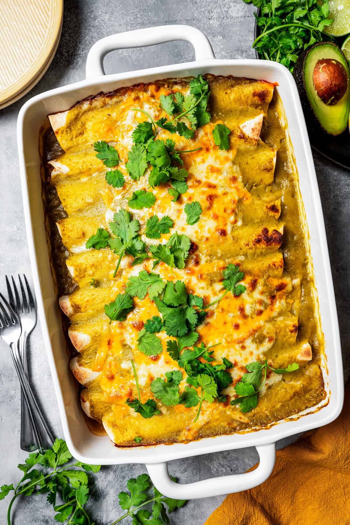 Green chili chicken enchiladas in a baking dish garnished with fresh cilantro leaves. There's an avocado, two forks, and an orange kitchen towel set around the baking dish.