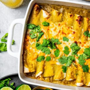 Overhead view of green chili chicken enchiladas in a baking dish garnished with fresh clantro leaves, next to bowls of lime halves and sliced jalapeños.