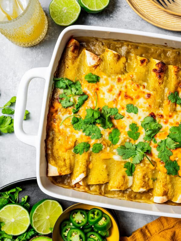 Overhead view of green chili chicken enchiladas in a baking dish garnished with fresh clantro leaves, next to bowls of lime halves and sliced jalapeños.
