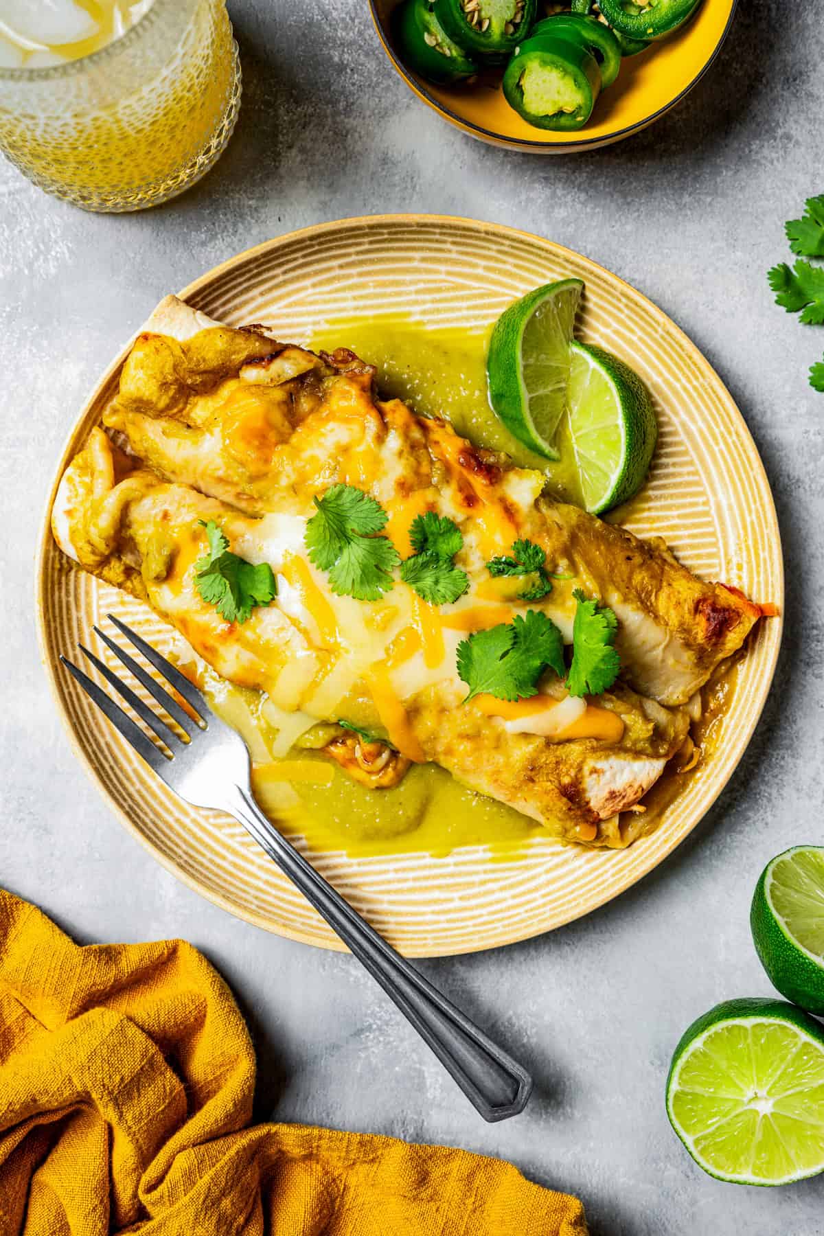 Two enchiladas are served on a dinner plate and garnished with cilantro. Next to the plate is a bowl of sliced jalapenos and a tall drinking glass.