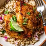 A bowl with chicken thigh served over a bed of rice with a garnish of lime wedges and green herbs.