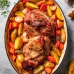 A whole pork shoulder roast is set in the center of a baking dish and is surrounded by sliced potatoes, carrots, and onions.