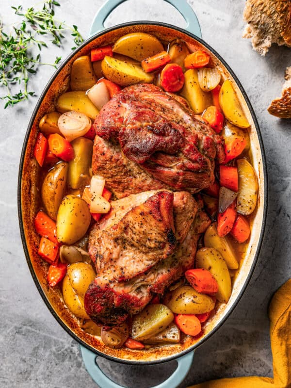 A whole pork shoulder roast is set in the center of a baking dish and is surrounded by sliced potatoes, carrots, and onions.