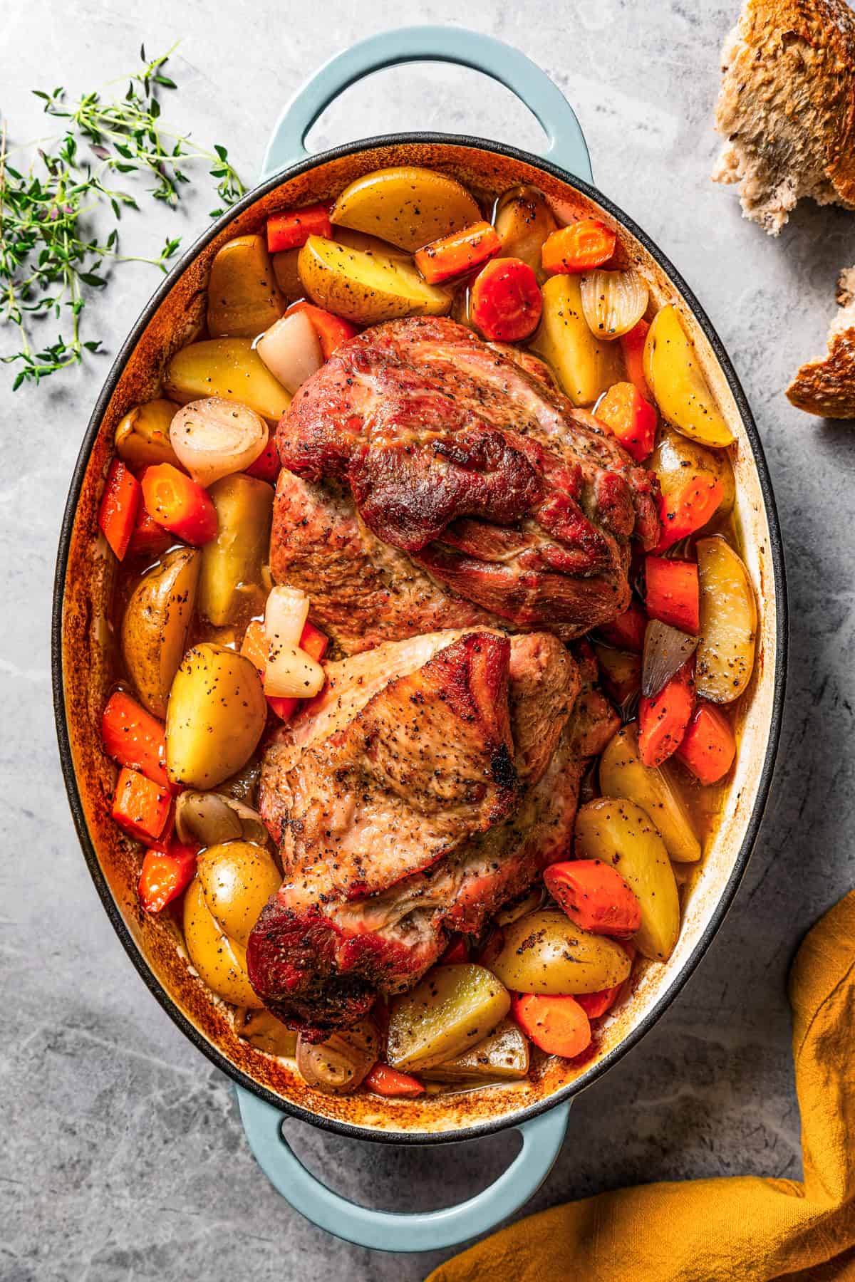 A whole pork shoulder roast is set in the center of a baking dish, surrounded by sliced potatoes, carrots, and shallots.