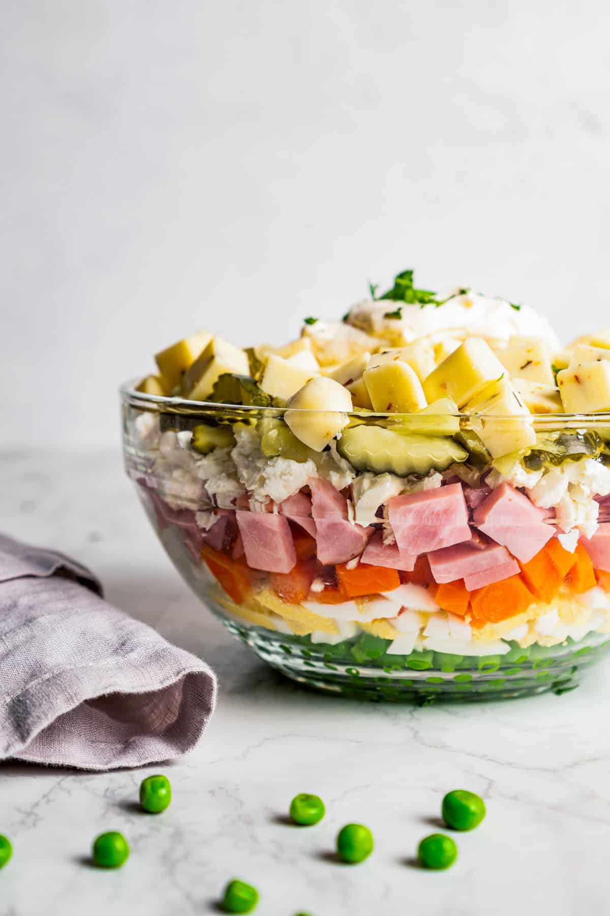 Side view of a layered Russian salad in a glass bowl next to a second bowl filled with peas, and peas scattered on the countertop.