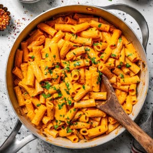 Overhead view of spicy rigatoni alla vodka garnished with chopped parsley in a large skillet, with a wooden spoon and small bowls of garnishes on the side.