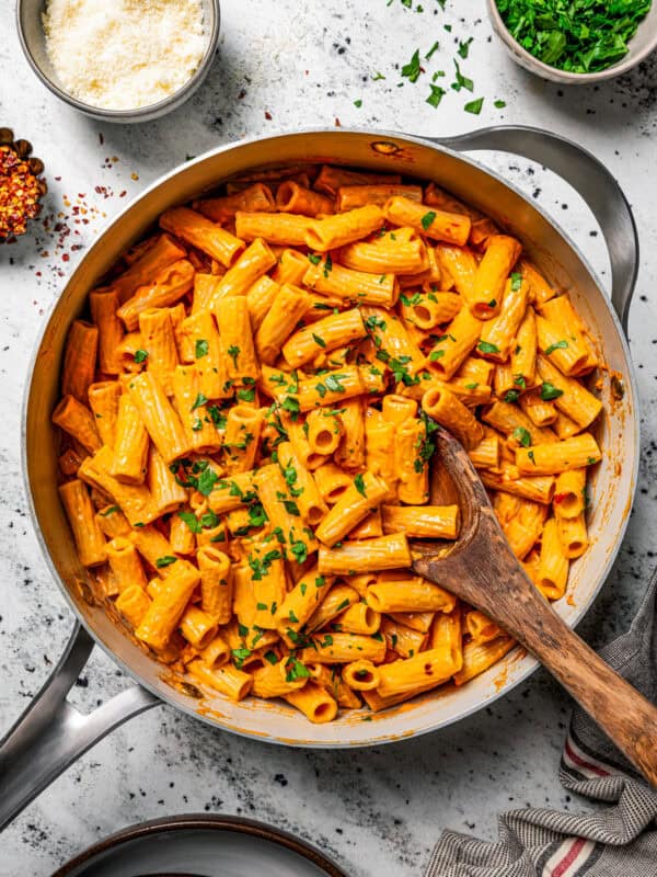 Overhead view of spicy rigatoni alla vodka garnished with chopped parsley in a large skillet, with a wooden spoon and small bowls of garnishes on the side.