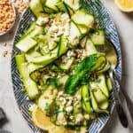 Overhead view of a salad made of zucchini ribbons and served on a large platter garnished with fresh basil leaves and lemon wedges, next to a set of salad tongs.