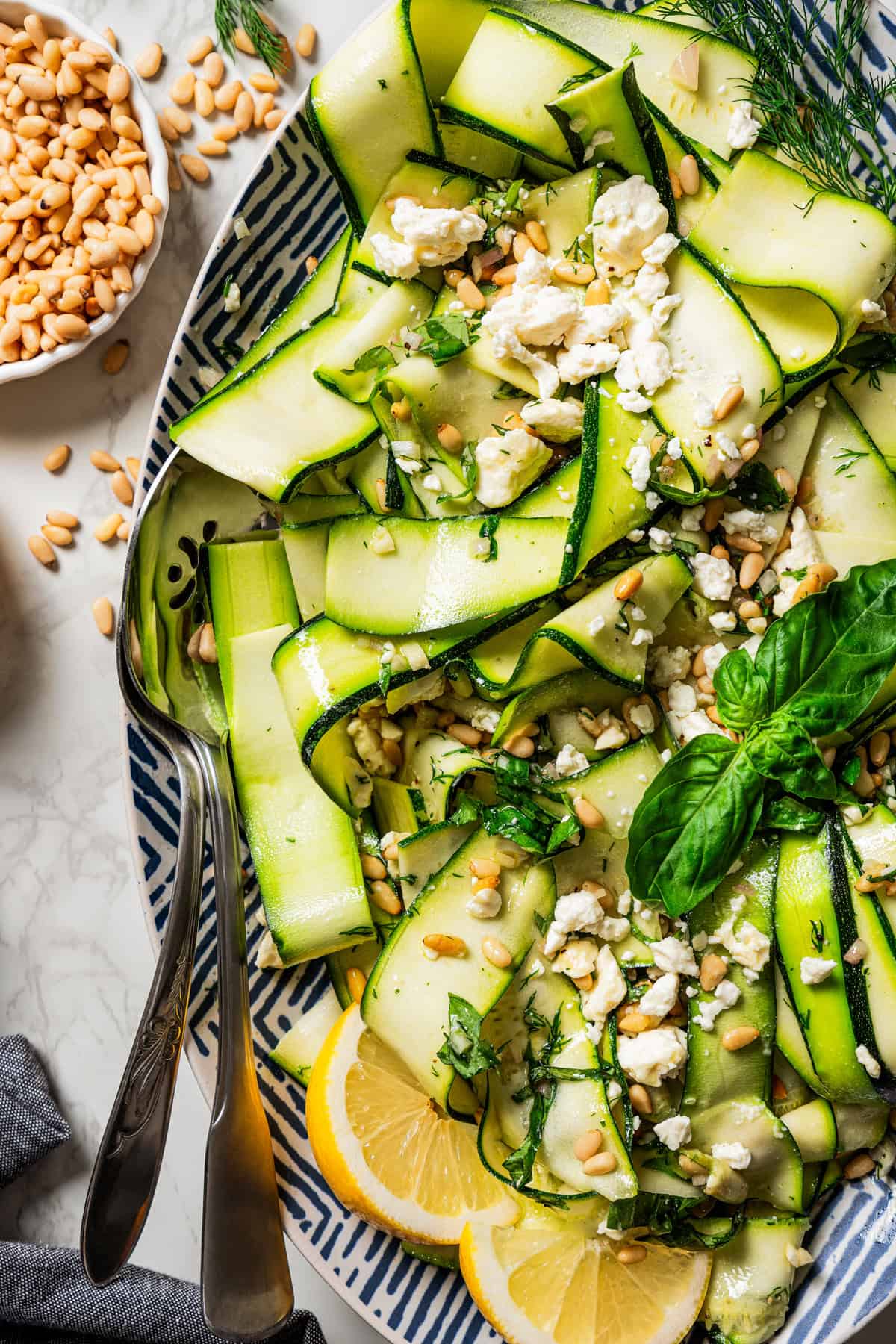 A plate of thinly sliced zucchini topped with pine nuts and crumbles of cheese, and a pair of salad tongs are set on the plate.