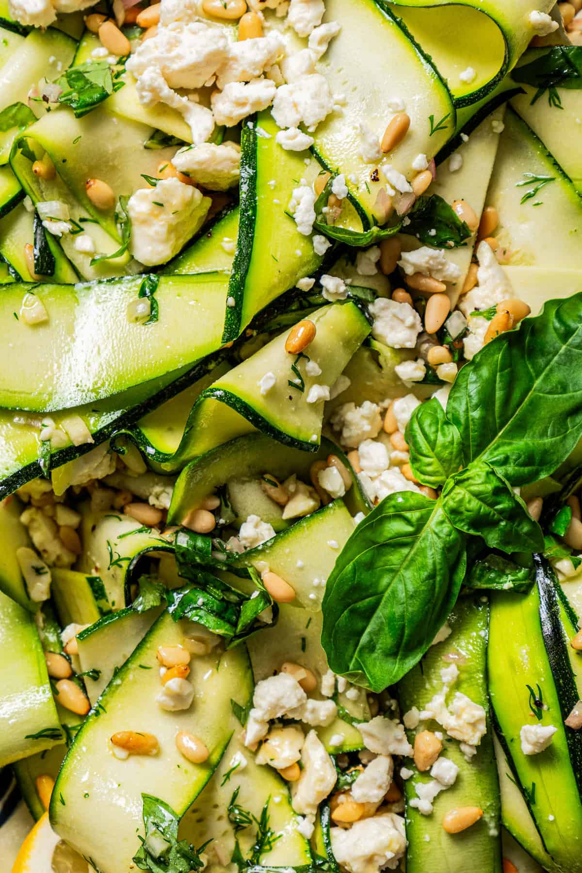Close-up image of a zucchini salad tossed with nuts, white crumbles of cheese, and fresh basil.