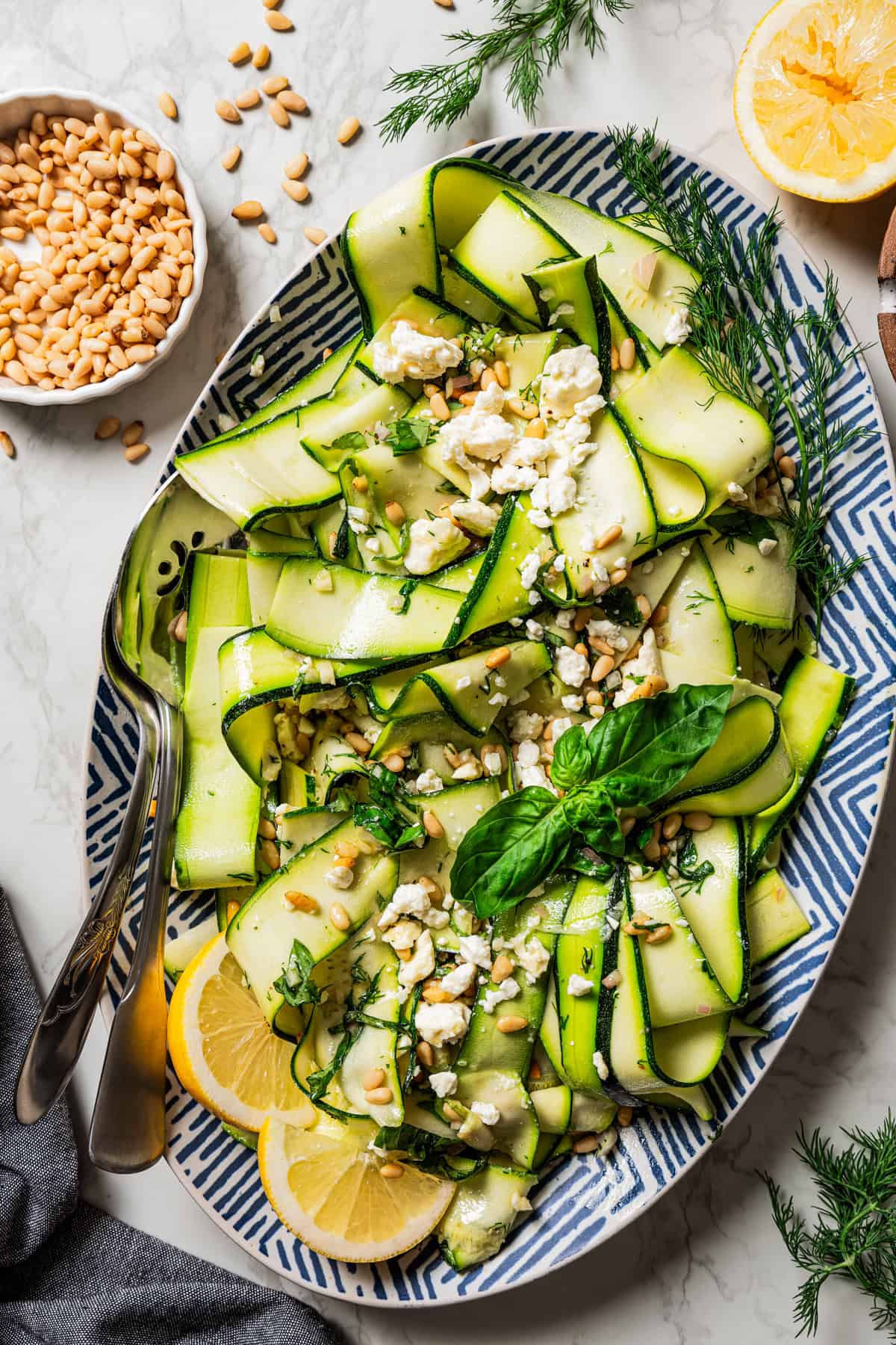 Overhead view of zucchini salad on a large platter garnished with fresh basil leaves and lemon wedges, next to a set of salad tongs and a bowl of toasted pine nuts.