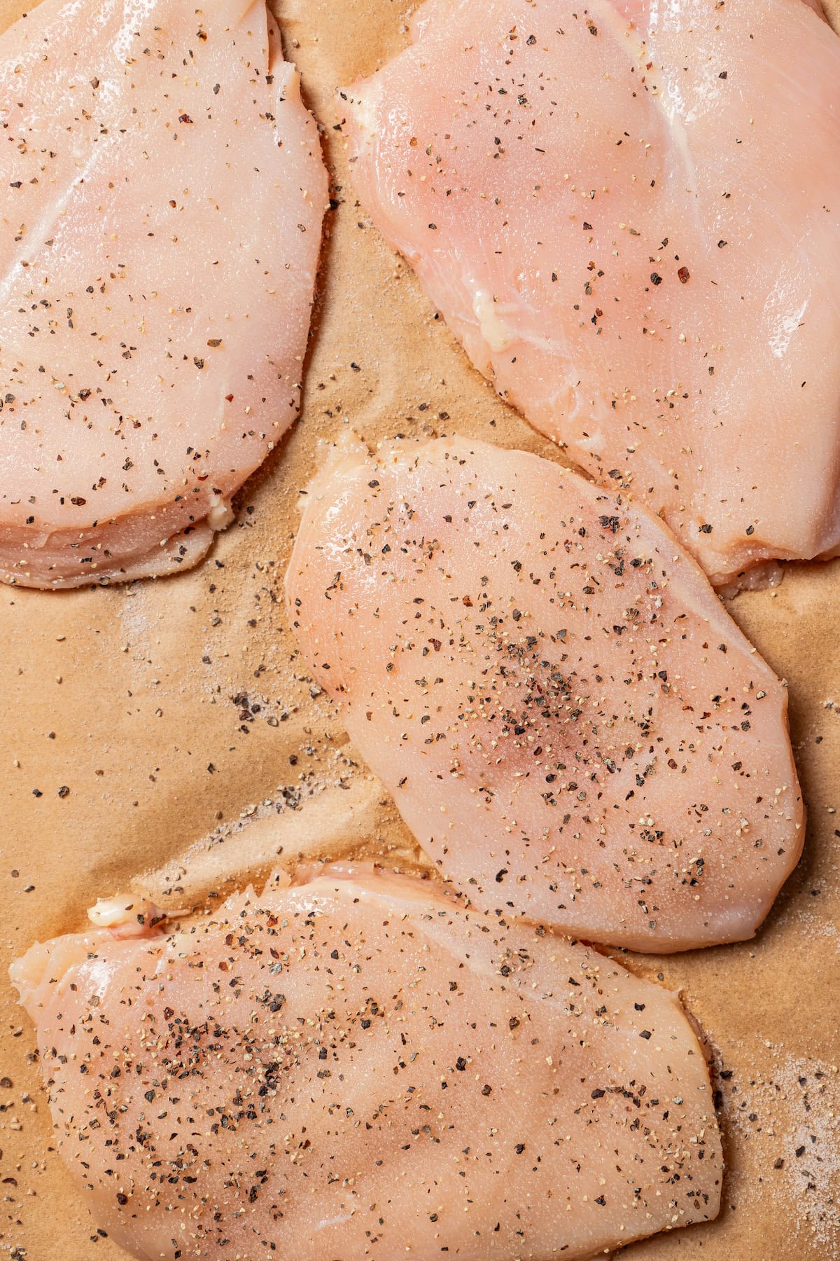 Uncooked chicken breasts pounded thin and seasoned with salt and pepper on a sheet of parchment paper.