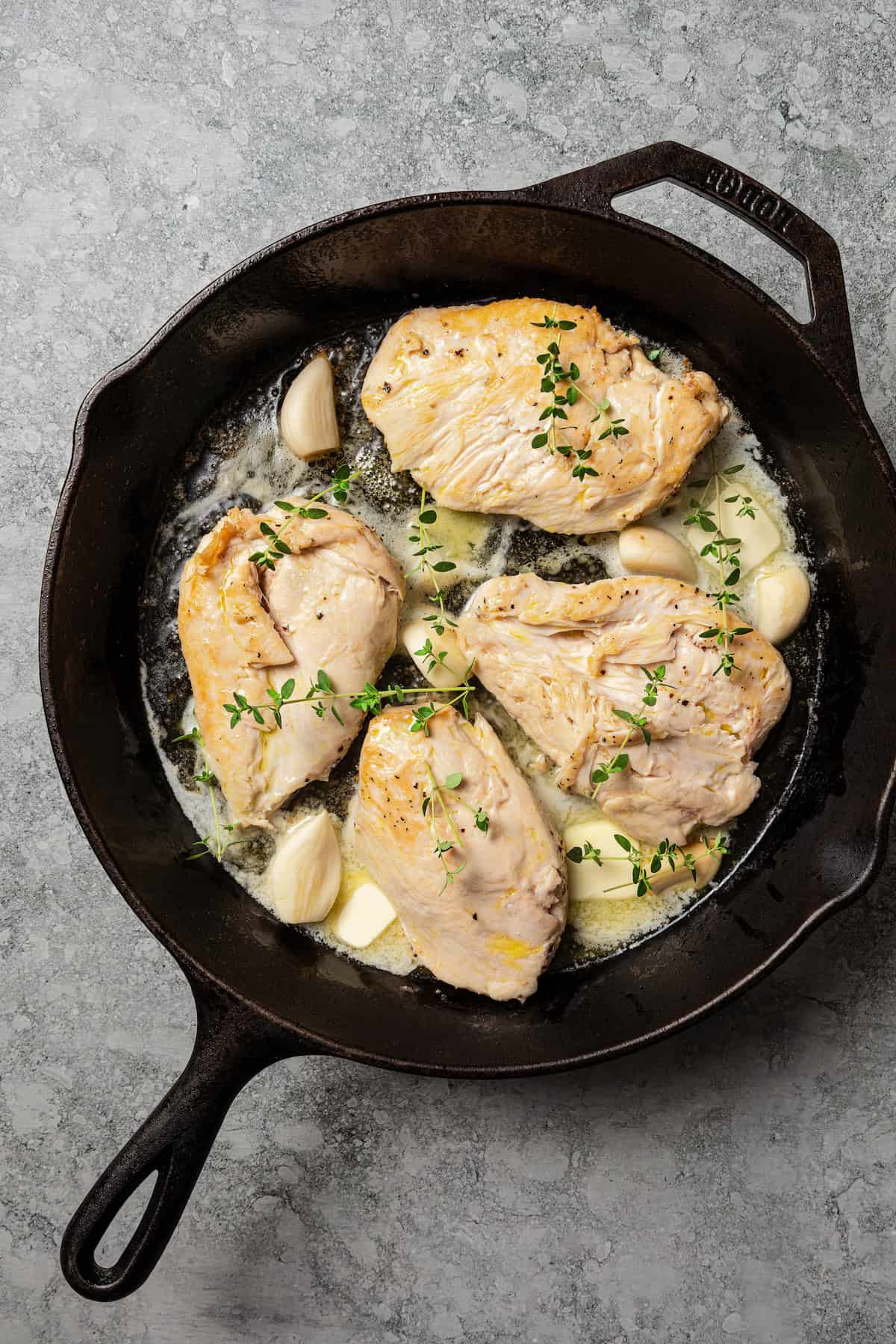 Four seasoned chicken breasts searing in a cast iron skillet with butter and herbs.