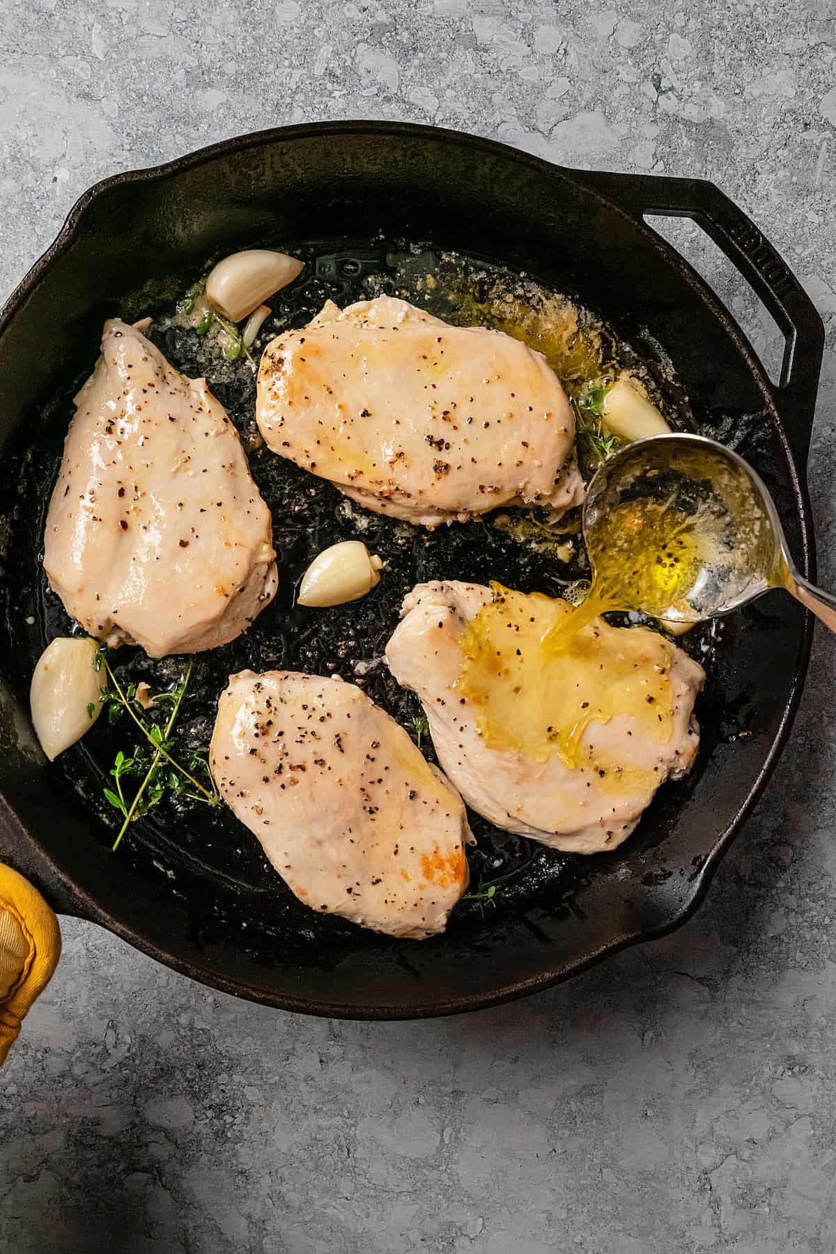 A ladle is used to baste chicken breasts with cooking liquid while they saute in a skillet.