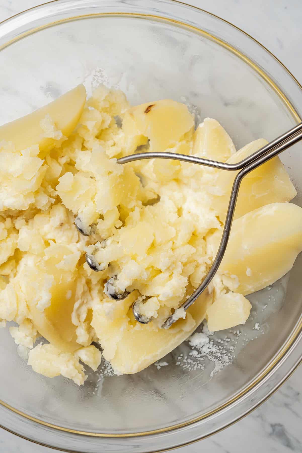 A potato masher resting in a glass bowl of partially mashed potatoes.