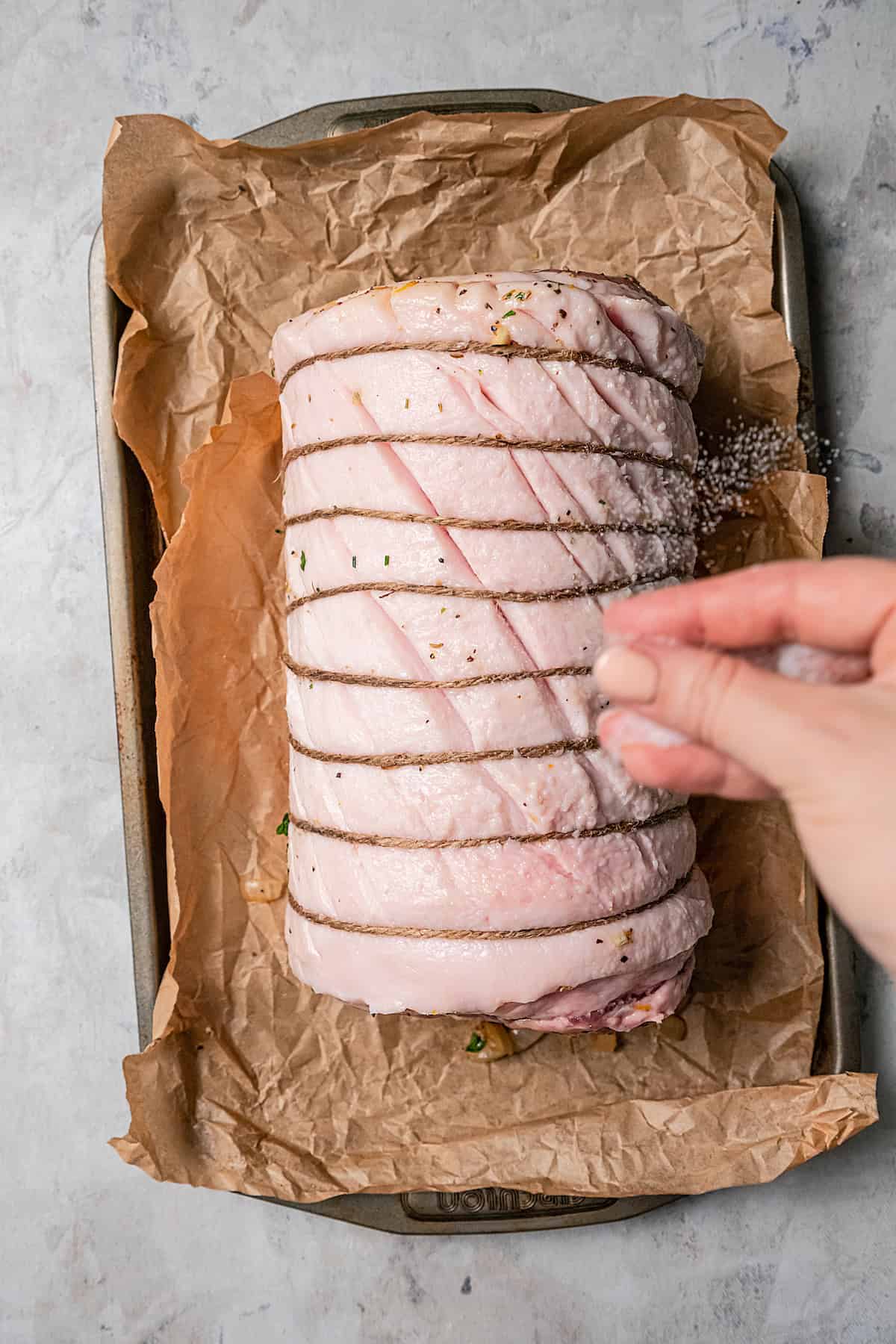 Overhead view of a hand sprinkling salt and pepper over rolled porchetta on a parchment-lined baking sheet.